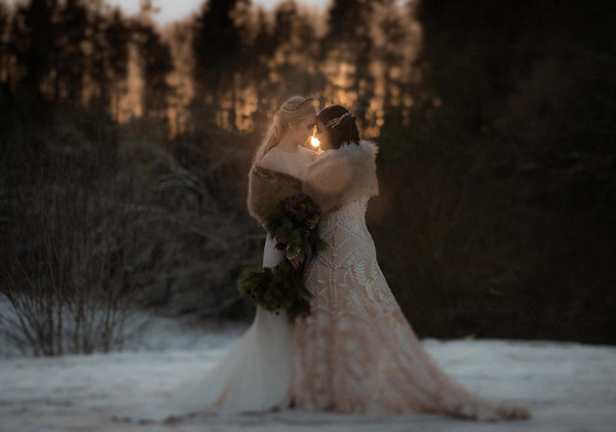 Two brides stand in snow wearing fur boleros embracing one another at sunset